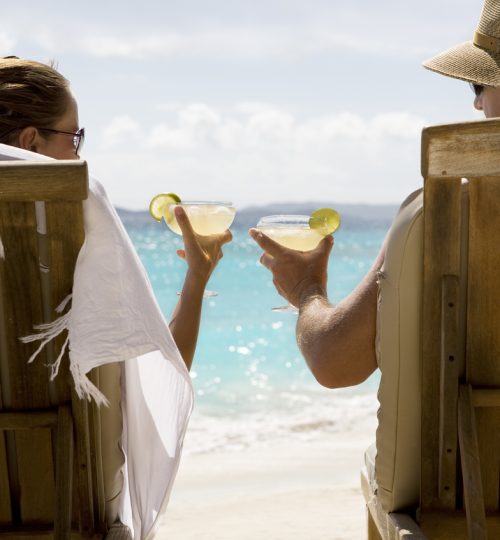 A man and woman sit in two wooden beach chairs facing the Caribbean ocean holding drinks and making a toast.